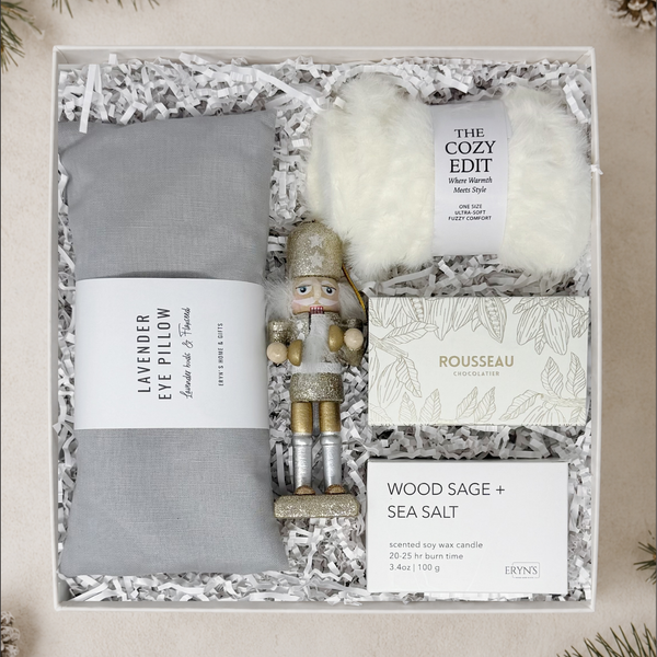 A curated gift box set, including a gray lavender eye pillow wrapped in a paper band, a white fluffy socks, a nutcracker figurine, a scented candle in a wood box, and a mini chocolate box.