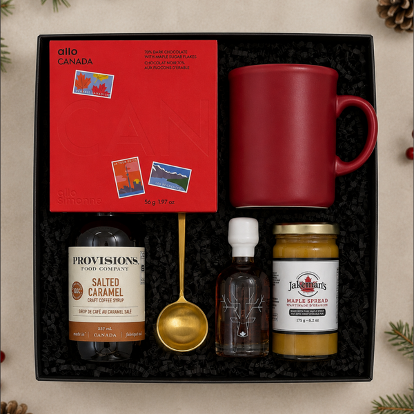 A curated gift box set containing a red ceramic mug, a red Allo Simonne chocolate bar, a bottle of salted caramel syrup, a jar of Escuminac's maple syrup, Jakeman's maple spread, and a gold spoon, all arranged in a black gift box on a neutral background with pine decorations.