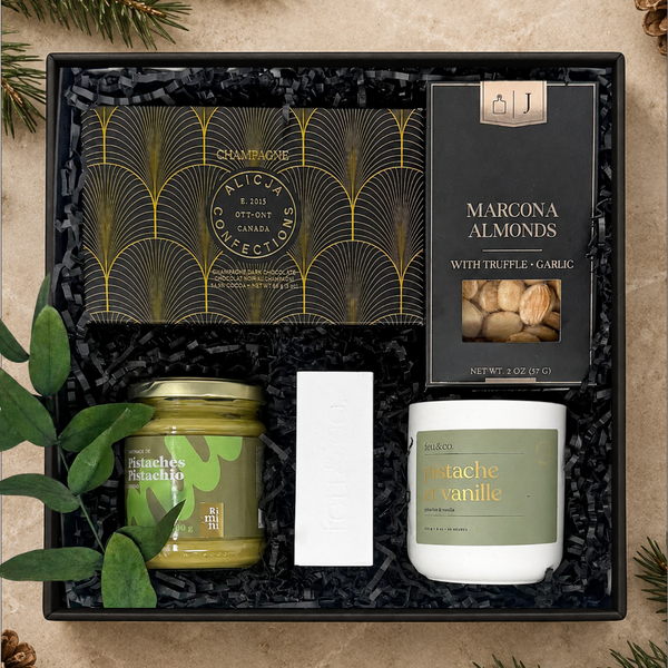 A curated gift box set arranged in a black box with black shredded filler. The set includes a Marcona Almonds box, a Champagne Chocolate bar, a jar of pistachio spread, a white candle, and a small white matches box.