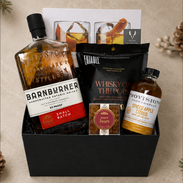 A curated gift basket or box containing a bottle of Barnburner whisky, a small bottle of Provision Spice Apple Citrus cocktail mix, a black bag of Eatable Whisky on the Pops popcorn, a small box of agave gummy bears, and two cocktail glasses, arranged in a black gift box.