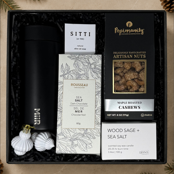 A curated gift box set containing a black insulated bottle, Sitti olive oil soap, Rousseau sea salt chocolate bar, Popinsanity maple roasted cashews, and a Wood Sage + Sea Salt scented wax candle, arranged in black crinkle paper inside a black gift box.