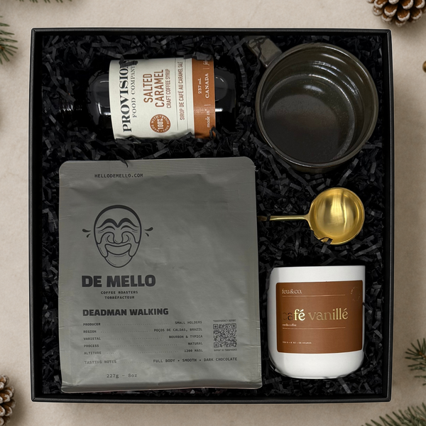 A curated coffee gift set arranged in a black presentation box on a light beige surface with pine branches and pinecones around it. The set includes a bottle of Provisions Salted Caramel coffee syrup, a bag of De Mello Deadman Walking coffee, a dark ceramic mug, a white candle labeled café vanillé, and a gold scoop.