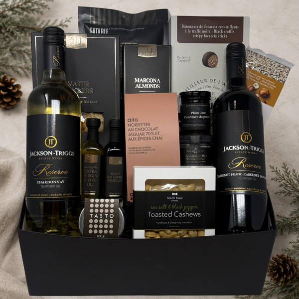 A curated gourmet gift arranged in a black rectangular box on a neutral fabric surface with pinecones and evergreen branches. The set includes two bottles of Jackson Triggs wine, olive oil and balsamic vinegar, focaccia sticks, crackers, rich chocolates, popcorn, balsamic pearls, jams, chilli salt, and nuts.