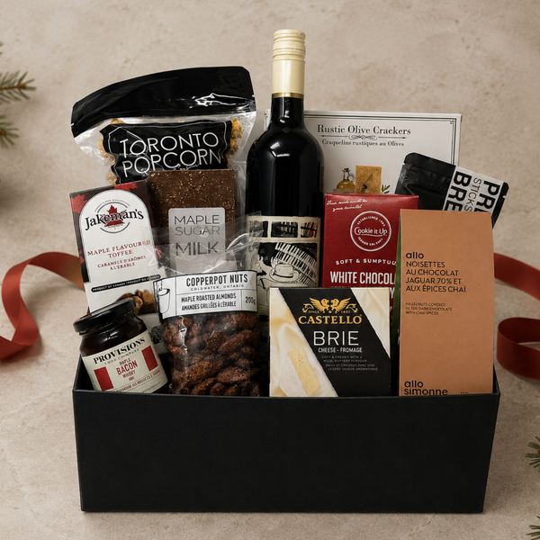 A gourmet gift basket arranged in a black rectangular gift box on a beige surface. The box includes a bottle of red wine, a bag of Toronto Popcorn, Castello Brie cheese, white chocolate, maple syrup, coffee nuts, and other snack items.